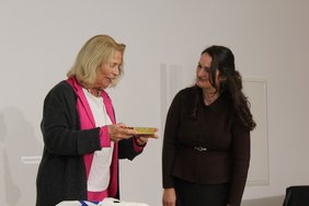 Rose-Lore Scholz (links), die stellvertretende Vorsitzende und Kulturbeauftragte des BdV-Landesverbandes Hessen, übergibt Dr. Violeta Bakia (rechts), der Leiterin des Hauses der Donauschwaben in Sindelfingen, als Geschenk eine Packung Ananastörtchen aus Wiesbaden. Bildnachweis: BdV-Landesverband Hessen