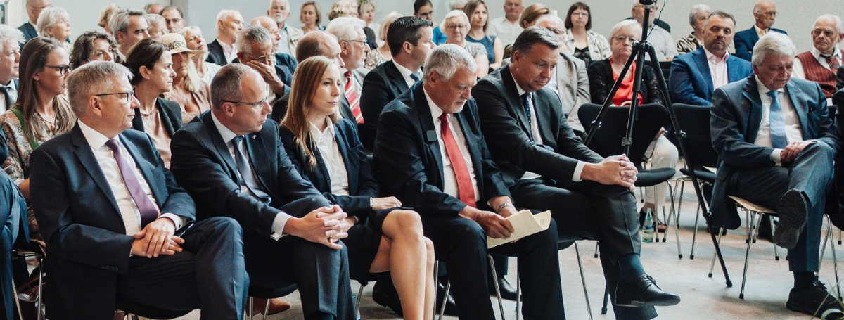 Ein große Gruppe von Personen die halbkreisförmig in Stuhlreihen sitzen alle festlich gekleidet